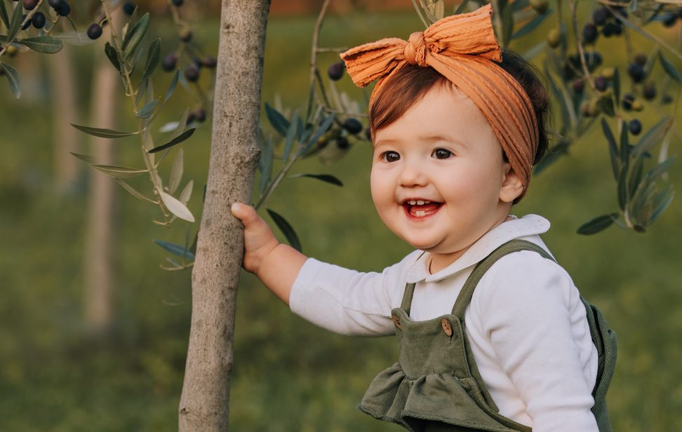 Bambina con un fiocco arancione che sorride appoggiata a un albero durante la sessione del primo compleanno